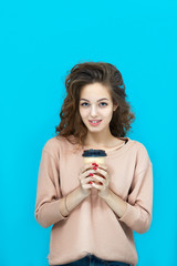 Portrait of a young beautiful woman wearing sweatshirt holding a cup with hot drink in hands and looking at you coquettishly over blue background