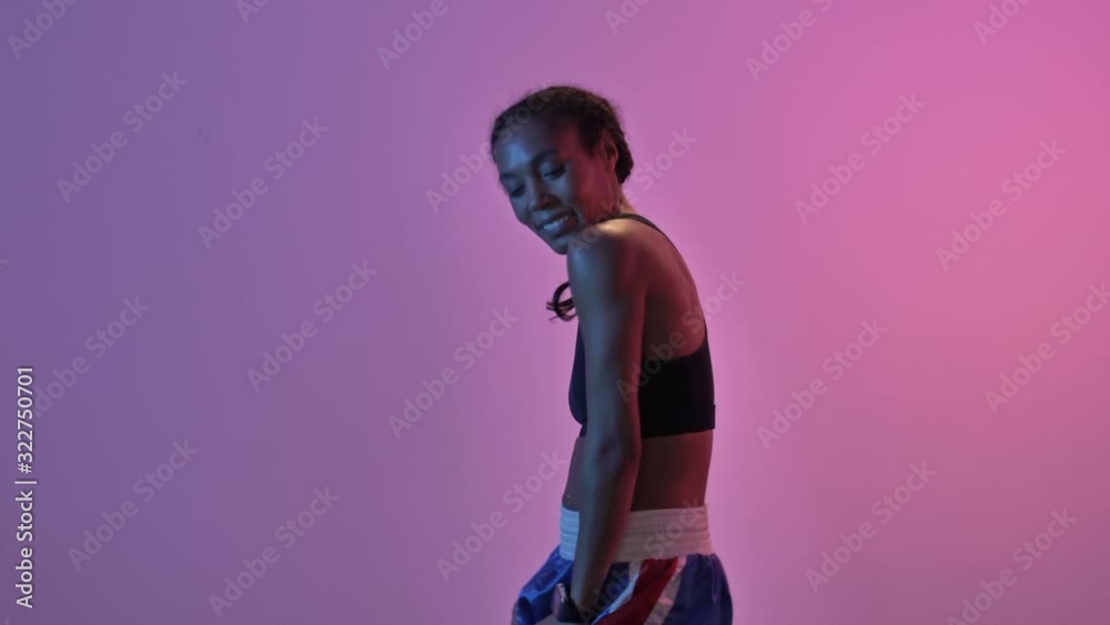 Wall mural Happy african sports woman dancing and looking at the camera isolated led flashlights background