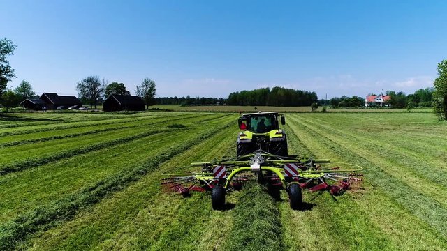 Drone aerial shot following a green tractor straight collecting grass on a Swedish summers field with sunny blue skies then tilting the camera down and flying above it.