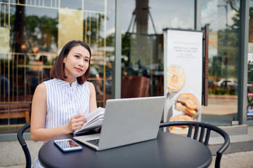 Beautiful confident woman in casual clothing reading book and drinking coffee while sitting in cafe with laptop and notebook