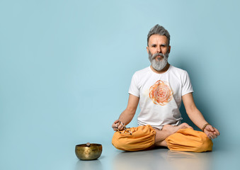 Elderly gray-haired man in dhoti clothes, holding rosary, sitting on floor in lotus pose on blue background. Singing bowl nearby - obrazy, fototapety, plakaty