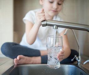 Little girl opens a water tap with her hand holding a transparent glass. Kitchen faucet. Glass of...