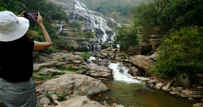 Young female tourist enjoying taking pictures of beautiful waterfall in northern Thailand.	