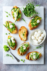 Appetizer crostini with mashed green pea, mozzarella, pea sprouts and zucchini ribbons on white marble board. Delicious healthy snack, spring appetizers