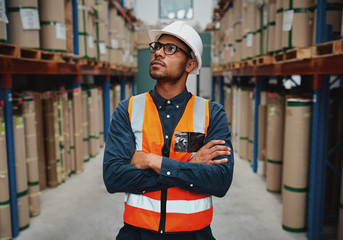 Confident warehouse supervisor in aisle standing with folded hands looking high at shelves with...