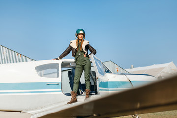 girl stands in a pose on the wing of an airplane in a helmet and a leather jacket with fur