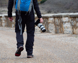 Man holding a camera with a big zoom objective outdoors in the middle of a path surrounded by a stone wall