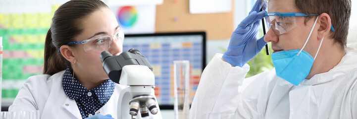 Portrait of laboratory co-workers investigating samples under microscope. Woman and man wearing...