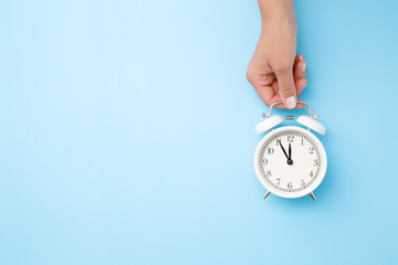 Woman hand holding white alarm clock on light blue table background. Pastel color. Time concept. Closeup. Empty place for text, quote or sayings. Top down view.