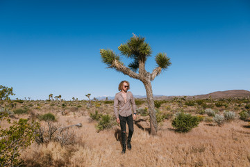 Joshua tree a curly haired blonde man is walking in the fierce heat and deep blue skies of the south western desert of North America, wearing dark sunglasses, a beige linen shirt