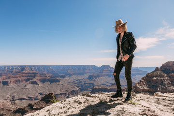 Fototapeta na wymiar A curly haired blonde man, wearing surprisingly a black leather jacket ,black ripped jeans, black shoes, and a beige linen shirt, is tipping his cowboy hat to the majestic beauty of the Grand Canyon