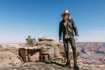 A curly haired blonde man, wearing surprisingly a black leather jacket ,black ripped jeans, black shoes, and a beige linen shirt, is tipping his cowboy hat to the majestic beauty of the Grand Canyon