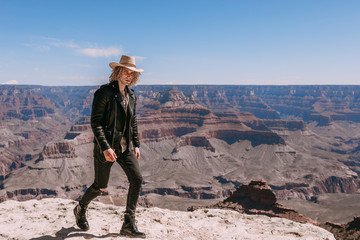 A Curly haired blonde man, wearing a black leather jacket ,black jeans, black shoes, beige linen shirt and matching cowboy hat, background vistas of the Grand Canyon