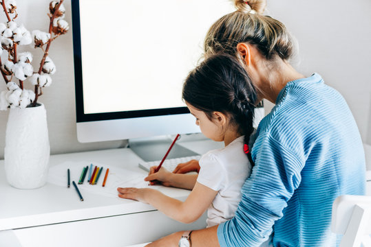 Rear view image of young woman with daughter sitting at home and working. Young mother with toddler child working at the computer and playing. Housewife mother sitting with her child and working.