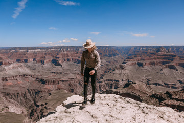 A Curly haired blonde man, wearing a beige T-Shirt ,black jeans, black shoes, beige linen shirt and matching cowboy hat, background vistas of the Grand Canyon