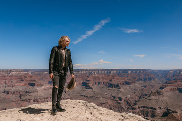 Fototapeta premium A Curly haired blonde man, wearing a black leather jacket ,black jeans, black shoes, beige linen shirt and matching cowboy hat, background vistas of the Grand Canyon
