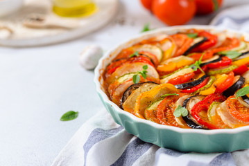 Ratatouille casserole on white background. Colorful layers of fresh summer vegetables: zucchini, eggplant, tomatoes and potatoes. Healthy vegan, vegetarian dish. Side view, copy space. Menu, recipe