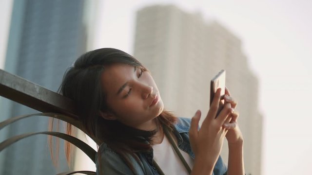 Asian women feel sad while sitting using a smartphone On a blurred building background in the sunset. Asia girl look mobile phone with lonely and alone