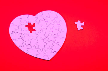 Pink jigsaw puzzle in the shape of a heart missing a piece sitting nearby on red background