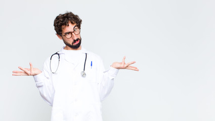 young doctor man shrugging with a dumb, crazy, confused, puzzled expression, feeling annoyed and...
