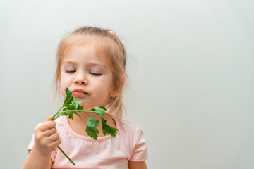 a little girl with a displeased look chews a bunch of parsley on a light smooth background. lack of vegetables in baby food