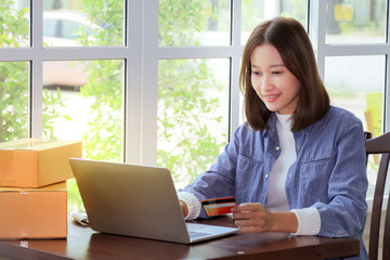 Beautiful Asian woman in casual using laptop technology for running small business start up marketing with cardboard box logistic delivery. young entrepreneur using credit card shopping payment,