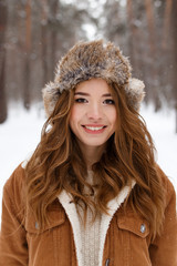 A beautiful cute cheerful young girl with curly hair and a snow-white smile in a brown jacket and fur hat walks and plays with snow in the winter forest against the background of trees, enjoy