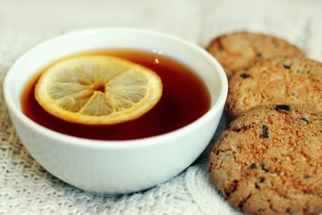 The cup of black tea with lemon and the oatmeal cookies on a white knitted background. Home comfort warms. Close-up