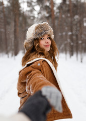 Fototapeta na wymiar A beautiful cute cheerful young girl with curly hair and a snow-white smile in a brown jacket and fur hat walks and plays with snow in the winter forest against the background of trees, enjoy