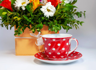 Red cup and saucer in white polka dots on the background of a multi-colored bouquet of flowers on a white table. Greeting card, mother's day