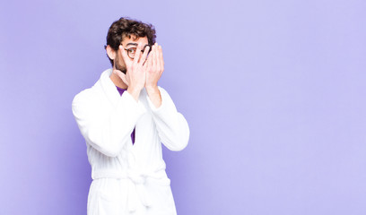 young bearded man wearing bathrobe feeling scared or embarrassed, peeking or spying with eyes...