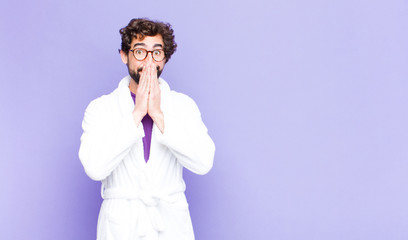 young bearded man wearing bathrobe happy and excited, surprised and amazed covering mouth with...