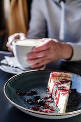Delicious cheesecake, with fresh berries on a plate, against the background of hands holding a cup of cappuccino, in natural light
