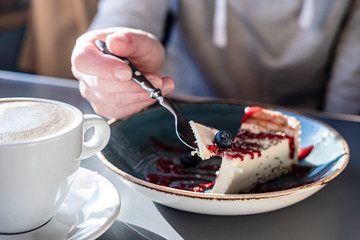 Cheesecake with fresh berry on a spoon, against background of a piece on a plate, next to a cup of cappuccino, in natural light.