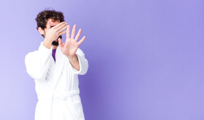 young bearded man wearing bathrobe covering face with hand and putting other hand up front to stop...