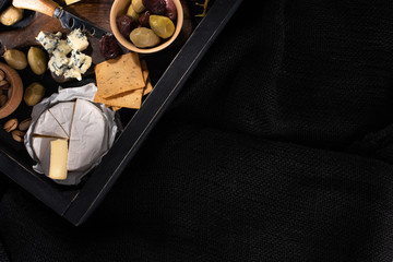 Top view of tray with pieces of camembert, dorblu, dried olives, crackers and pistachios isolated on black