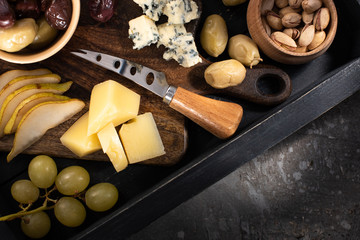 Top view of tray with pieces of cheese, dried olives, pistachios, slices of pear, grapes and knife on grey background