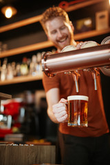 Handsome caucasian barman pouring beer while standing in pub. Selective focus on hand.