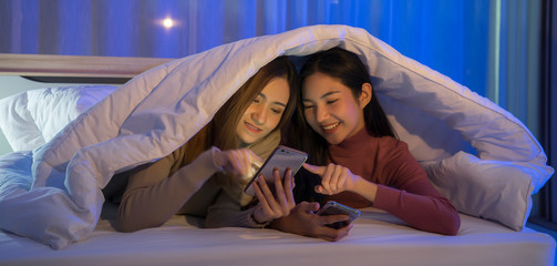 two young college female student roommates lying in bed under the quilt having fun laughing cheerfully. charming asian girls in pajamas nightwear smiling relaxing together in bedroom dormitory.