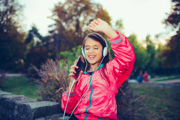 Close up portrait happy little girl dancing, singing and listening music with headphones and smart phone outdoor