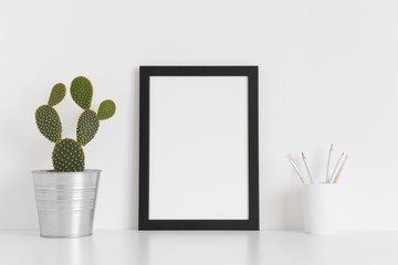 Black frame mockup with a cacutus in a pot and workspace accessories on white table. Portrait orientation.