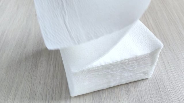 Stack of white square napkins on wooden table. Female hand takes one paper towel from above with fingers. Close-up, daylight