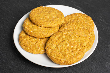 Group of six whole sweet golden oat cookie on white ceramic plate on grey stone