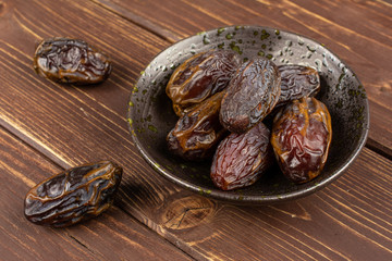 Lot of whole dry brown date fruit in dark ceramic bowl on brown wood