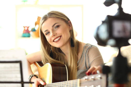 Happy Guitar Performer Recording Musical Vlog. Caucasian Cute Woman Sitting and Playing Music. Joyful Blogger Filming on Camera at Apartment. Cute Girl Bass Player Broadcast Rehearsal