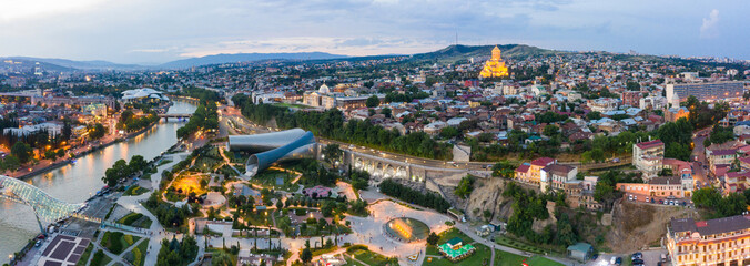 Panorama of the old town in the old district of Avlabari, Holy Trinity Cathedral and Rike Park, the Kura river reflects the evening city lights and cars traffic with blure in Tbilisi, Georgia © miklyxa