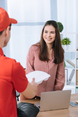 Selective focus of courier in red uniform giving food container to smiling businesswoman at table