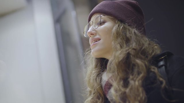 Low angle view of pretty woman with curly hair wearing trendy hat and scarf standing at shop window in downtown