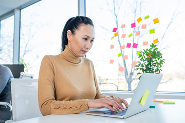 An Asian woman using a lap top in an office. She is typing on the lap top, there are sticky notes...