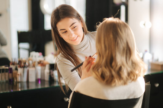 Beautiful female makeup artist doing makeup for a young blond hair girl in a beauty salon sitting in front of a large mirror. Concept of preparation for the holiday and meeting. Start working
